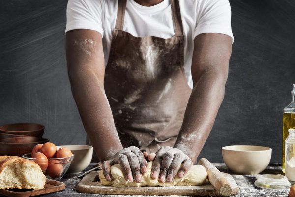 Cropped shot of dark skinned talented male cook kneads dough on wooden counter, going to bake delicious pie, uses different ingredients, going to treat guests. People, baking, culinary concept